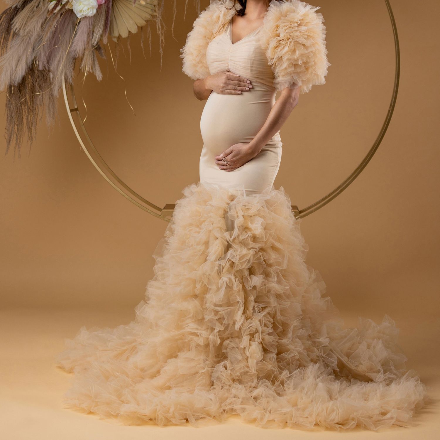 Pregnant woman in elegant champagne ruffle mermaid gown posing in front of gold circular arch backdrop.