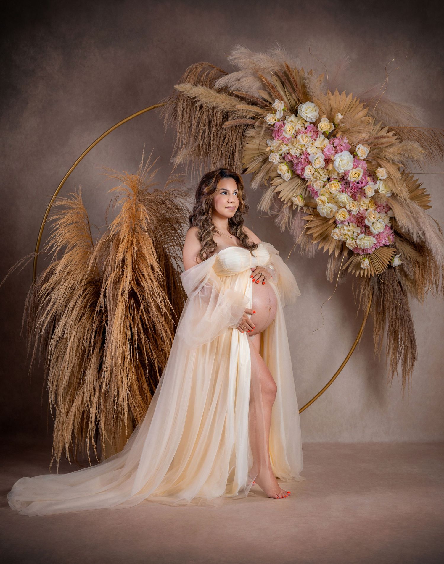 Pregnant woman in sheer champagne gown poses by floral hoop arch with pampas grass in elegant maternity studio photo.
