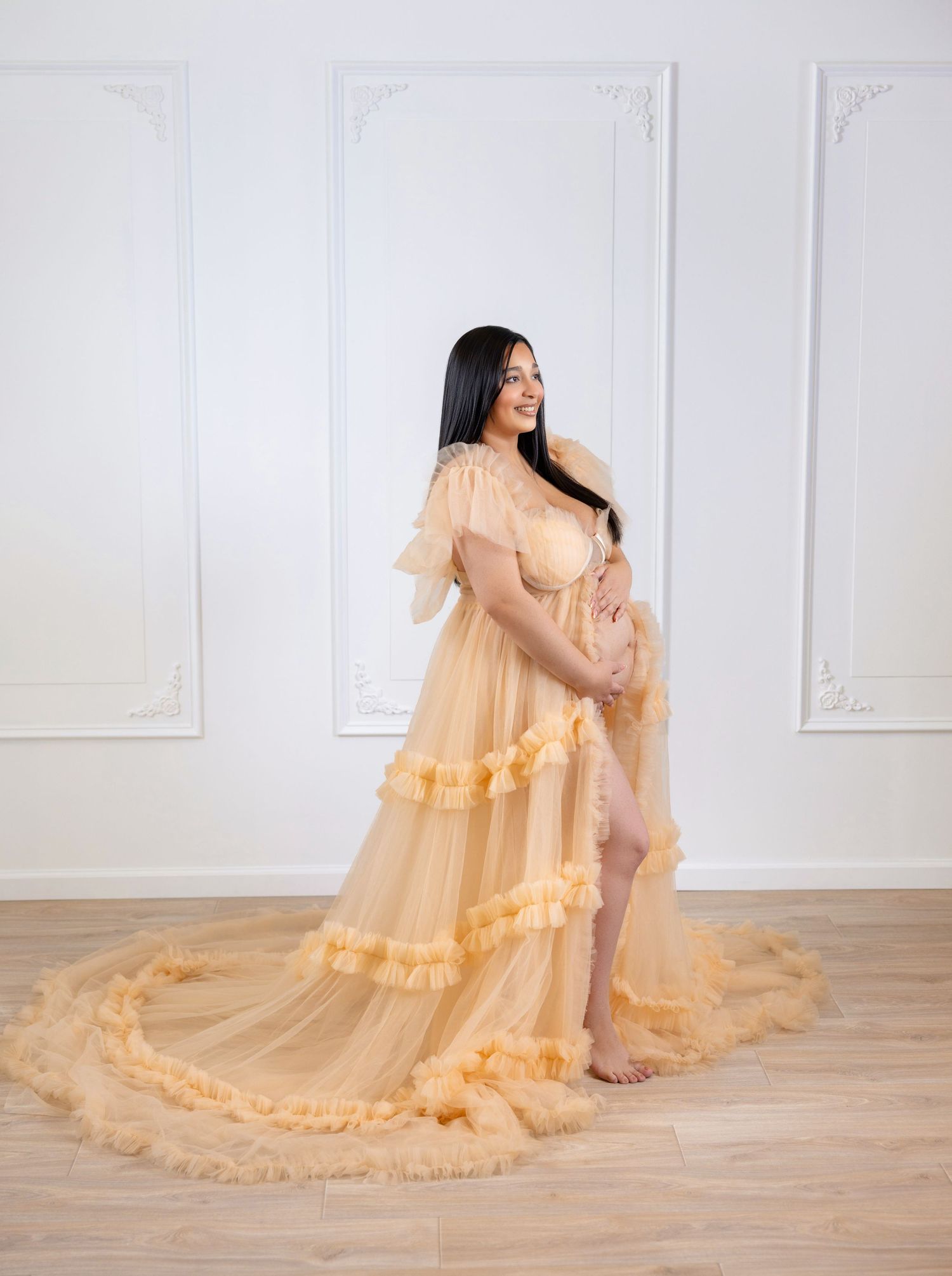 Pregnant woman in elegant peach tulle maternity gown with ruffles and train, posing in bright white studio.