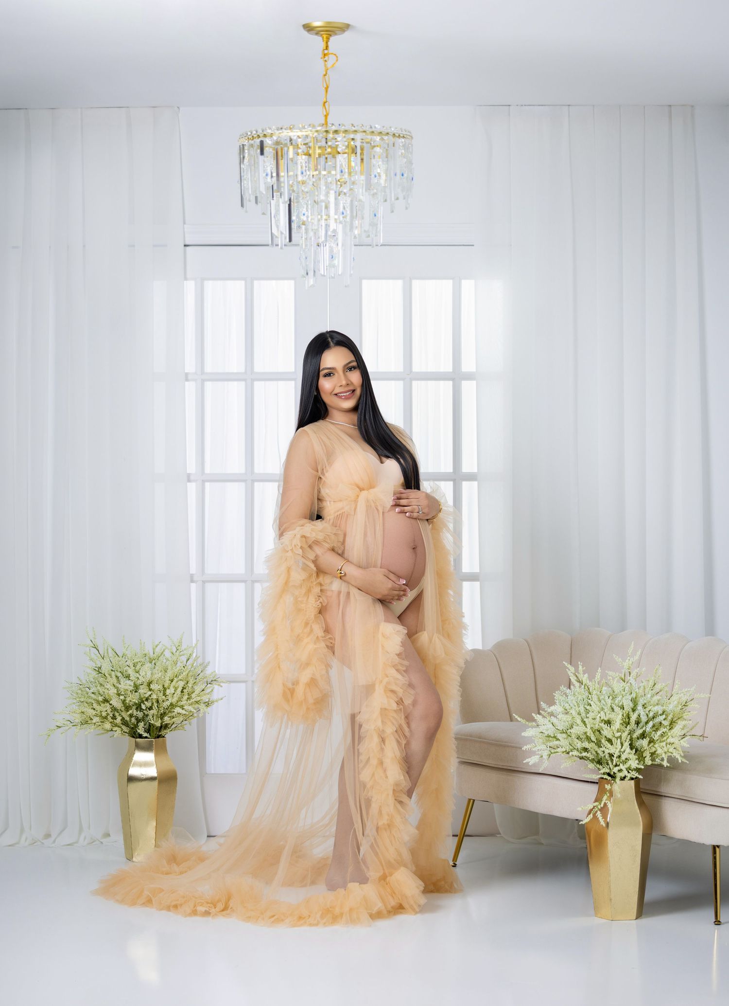Pregnant woman in elegant peach tulle maternity gown posing in bright studio with chandelier and gold vases.