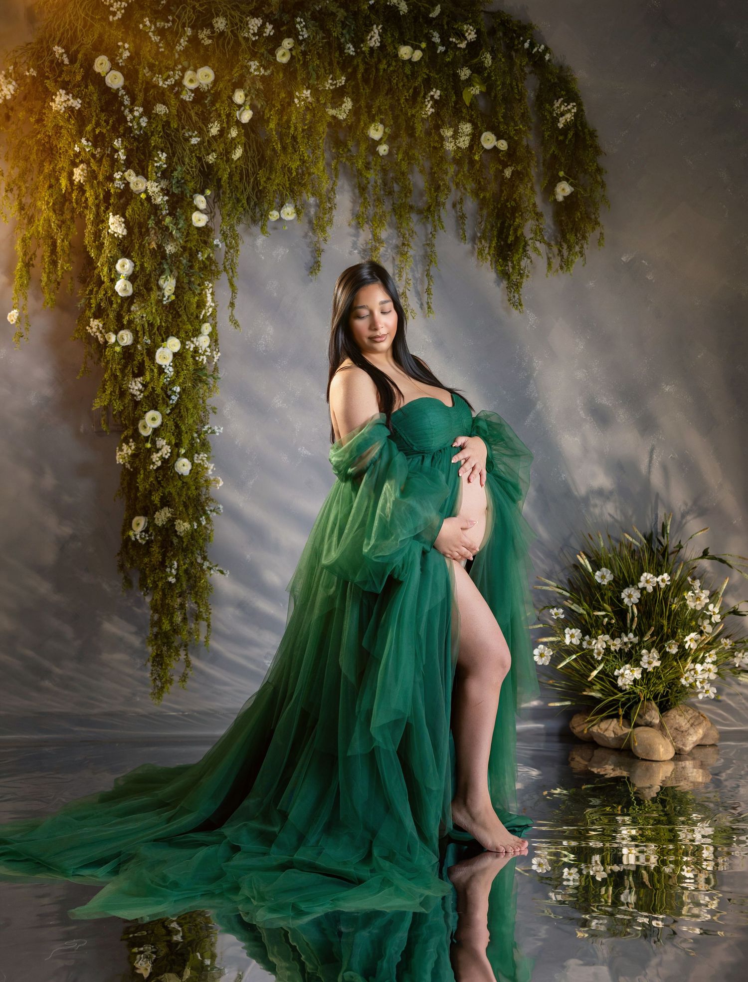 Pregnant woman in elegant green tulle gown poses among hanging flowers and greenery in a dreamy maternity portrait.