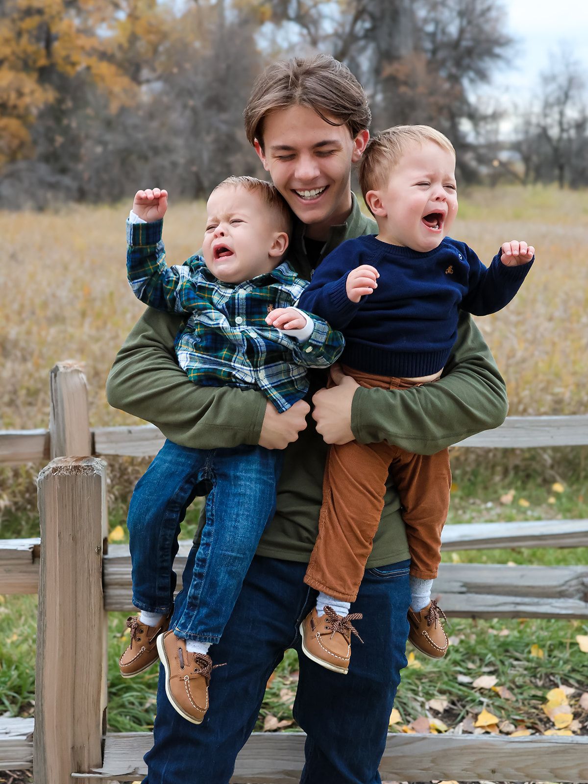 Smiling young adult holding two laughing toddlers outdoors near a wooden fence with autumn foliage in background.