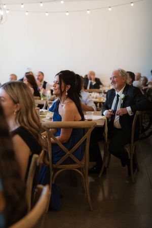 Wedding reception guests in formal attire seated at wooden tables under string lights in a bright venue.