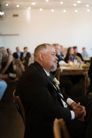 Older man in black tuxedo with boutonniere sits attentively at a wedding reception under string lights.