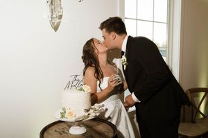 Bride and groom share a kiss beside their wedding cake at an elegant indoor reception venue.
