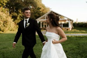 Bride and groom walk hand in hand across green lawn with white pavilion backdrop.