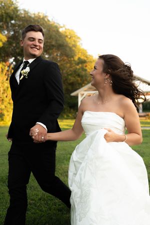 Bride and groom laugh joyfully while holding hands and running across green lawn at outdoor wedding.