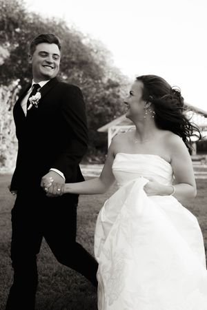 Black and white photo of a smiling bride and groom holding hands outdoors on their wedding day.
