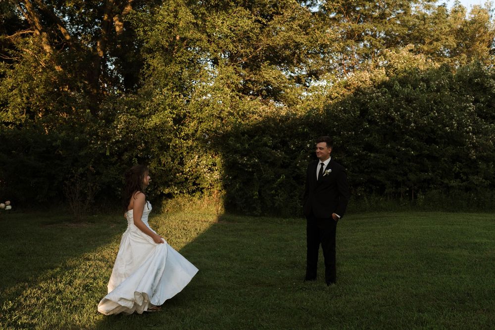 Bride in white gown and groom in black tux share a first look moment on a sunlit green lawn surrounded by trees.