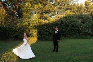 Bride in white strapless gown twirls on green lawn while groom in black suit watches during outdoor wedding photos.