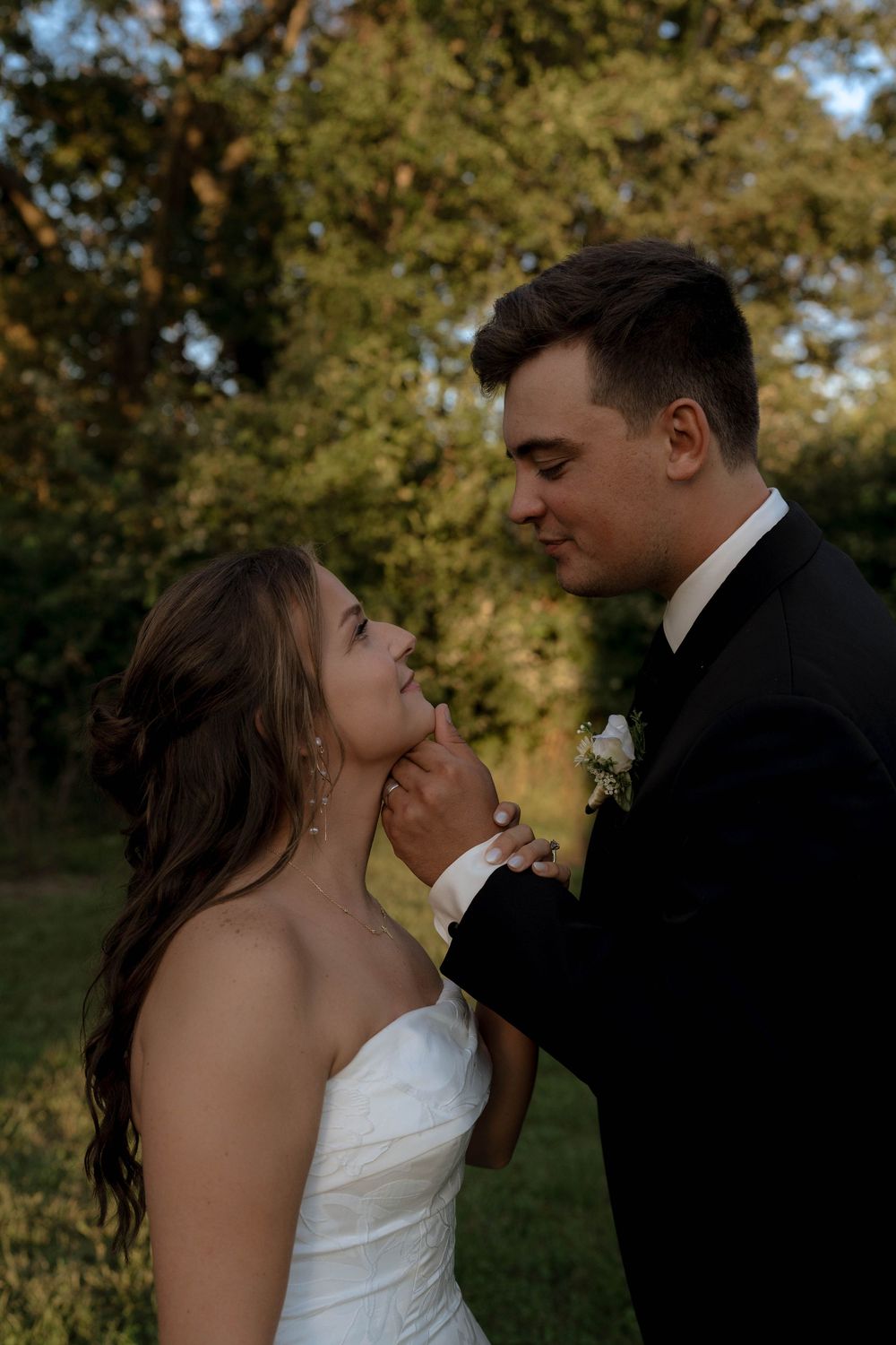 Groom tenderly holds bride's face as they gaze at each other outdoors at golden hour.