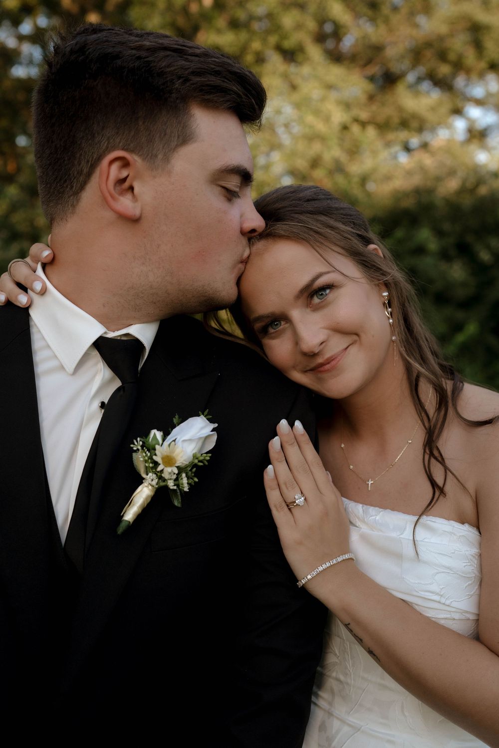 Groom in black suit kisses bride on cheek as she smiles, showing her diamond ring and white gown outdoors.
