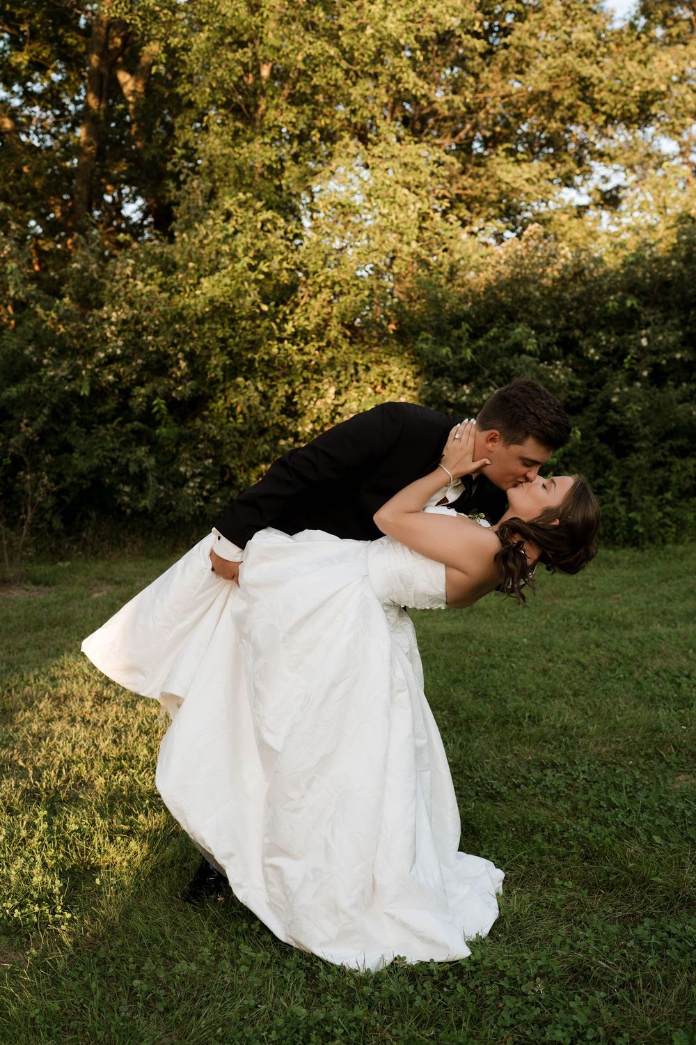 Groom dips bride for a romantic kiss in a lush green outdoor garden during their wedding celebration.