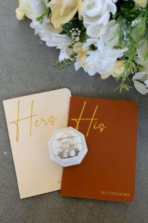 His and Hers wedding vow books with gold lettering, diamond engagement ring and wedding bands in white box, white floral bouquet.