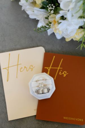 Wedding rings in velvet box on His and Hers vow books with white floral bouquet, elegant bridal detail photography.