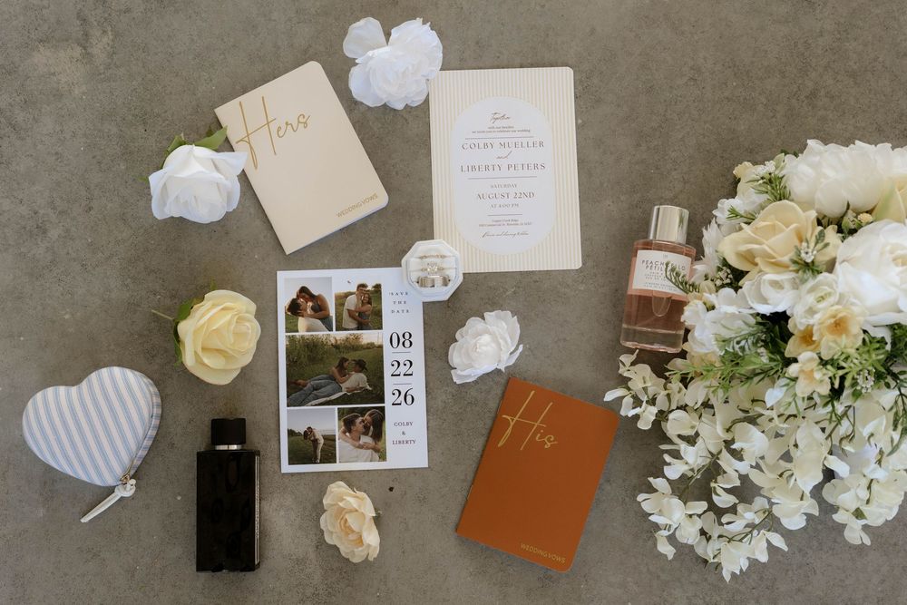 Elegant wedding flat lay with invitation suite, perfume, white florals, heart trinket box, and brown journal on gray surface.