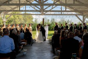 Bride and groom share a kiss at an outdoor wedding ceremony under a white pavilion with guests seated on both sides.