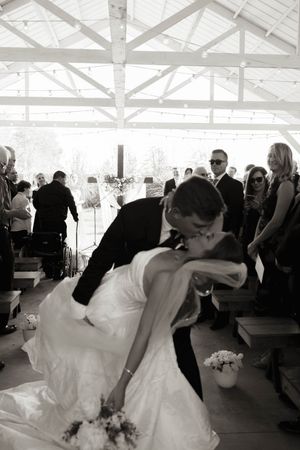 Black and white photo of bride and groom sharing a romantic dip kiss at wedding ceremony aisle.