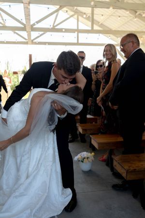 Groom dips bride for romantic kiss at wedding ceremony while guests watch and celebrate in rustic venue.