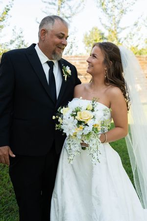 Bride in white strapless gown holding yellow and white bouquet smiles at man in black suit at outdoor wedding.