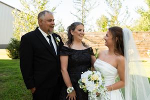 Bride in white gown with veil smiles alongside two family members outdoors at her wedding celebration.