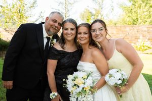 Family of four poses outdoors at wedding, bride in white gown holding bouquet with parents and sister.