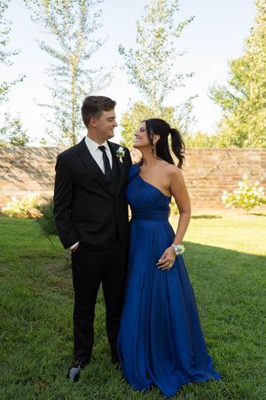 Groomsman in black suit with bridesmaid in blue one-shoulder gown smiling outdoors.