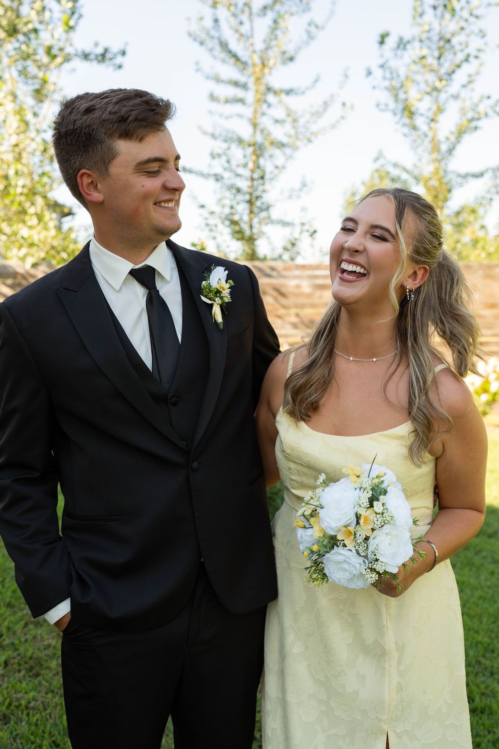 Smiling groomsman and laughing bridesmaid in yellow dress holding white bouquet outdoors.
