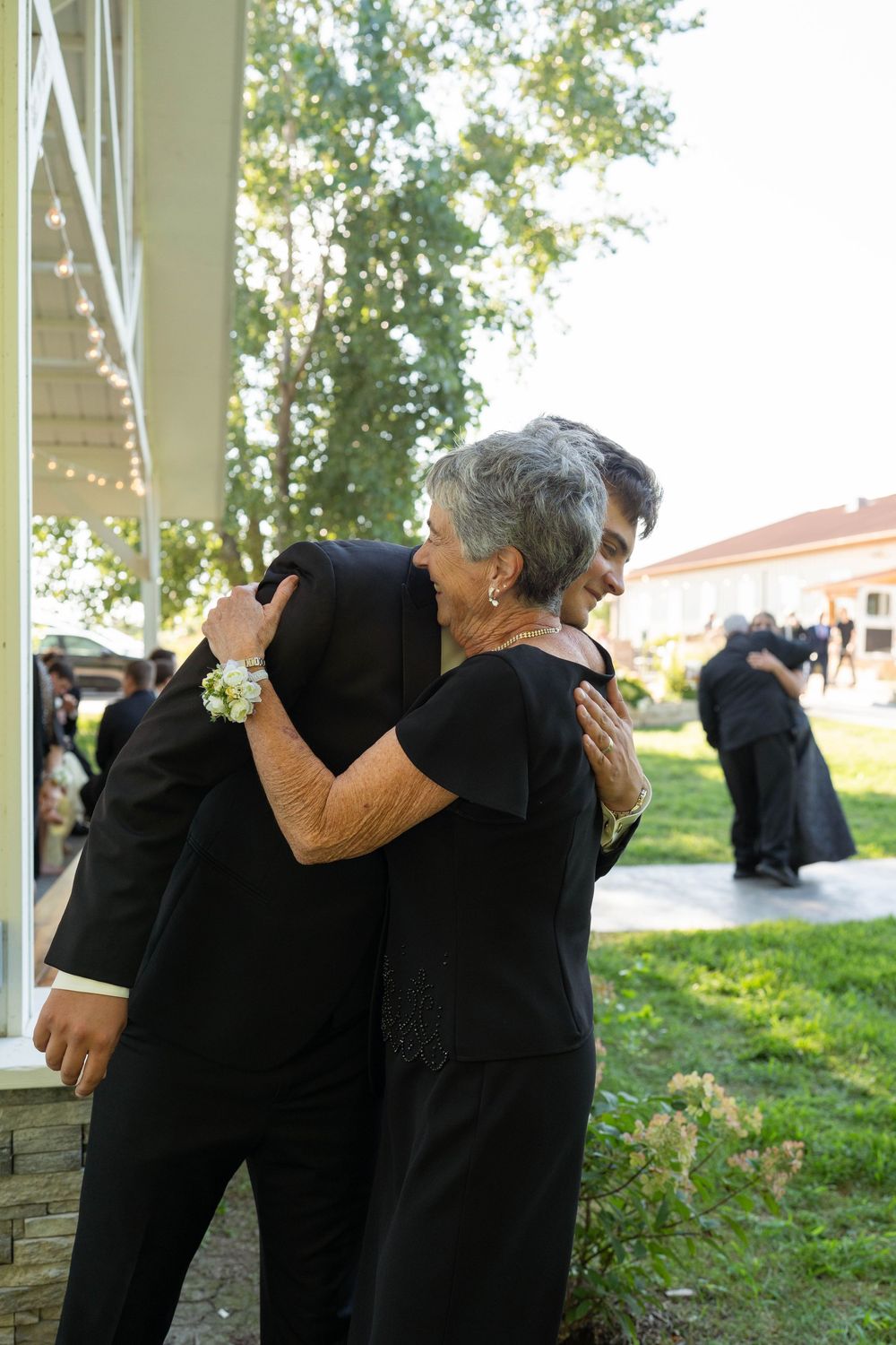 Groom hugging his mother outdoors at a wedding reception near a white building.