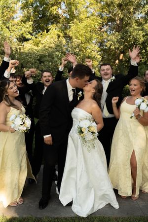 Bride and groom share a kiss surrounded by cheering wedding party in yellow dresses and black tuxedos outdoors.