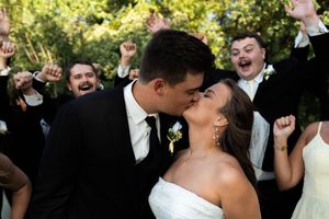 Bride and groom share a kiss surrounded by cheering groomsmen in black suits at their wedding.