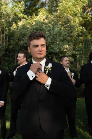 Handsome groom in black tuxedo adjusting tie with groomsmen standing behind him outdoors.