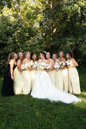 Bride in white gown surrounded by bridesmaids in yellow dresses holding white bouquets outdoors.
