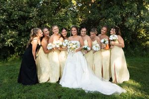 Bride poses with seven bridesmaids in gold dresses holding white bouquets under green trees outdoors.