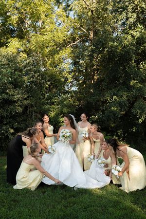 Bride in white gown surrounded by bridesmaids in yellow dresses in lush garden setting.