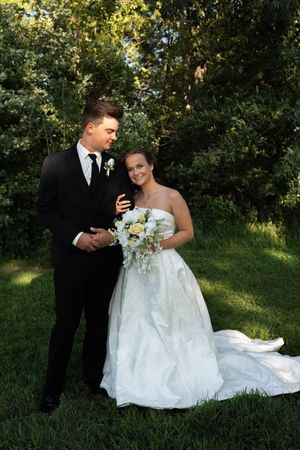 A bride in a white gown holding a bouquet stands with a groom in a black tuxedo on a lush green lawn.