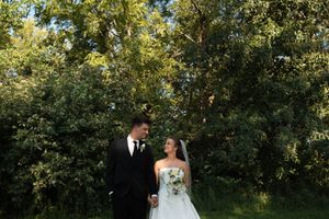 Bride in white strapless gown and groom in black suit walk hand in hand before lush green trees.