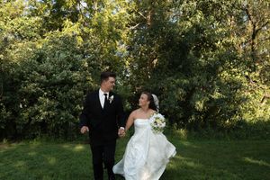 Bride in white gown holding bouquet walks hand in hand with groom in black suit through lush greenery.