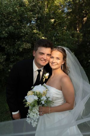 Smiling bride and groom pose together outdoors, bride holding white bouquet wearing veil, groom in black suit with boutonniere.