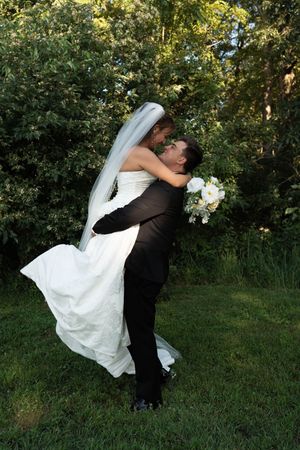 Groom lifting bride in white wedding dress for romantic kiss in green outdoor garden.
