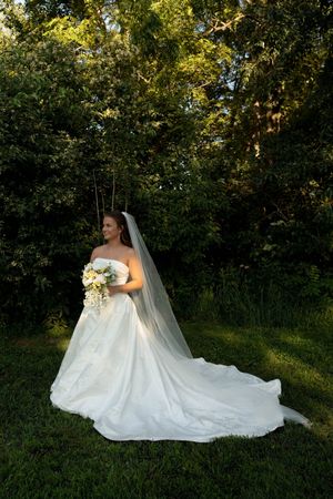 Bride in white strapless gown with long veil holds bouquet outdoors among lush green trees on her wedding day.