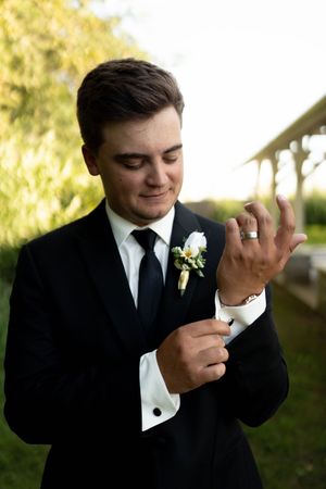Groom in black tuxedo adjusting cufflinks outdoors, wearing wedding ring and white boutonniere.