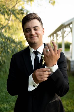 Young groomsman in black tuxedo smiling while showing wedding ring outdoors.