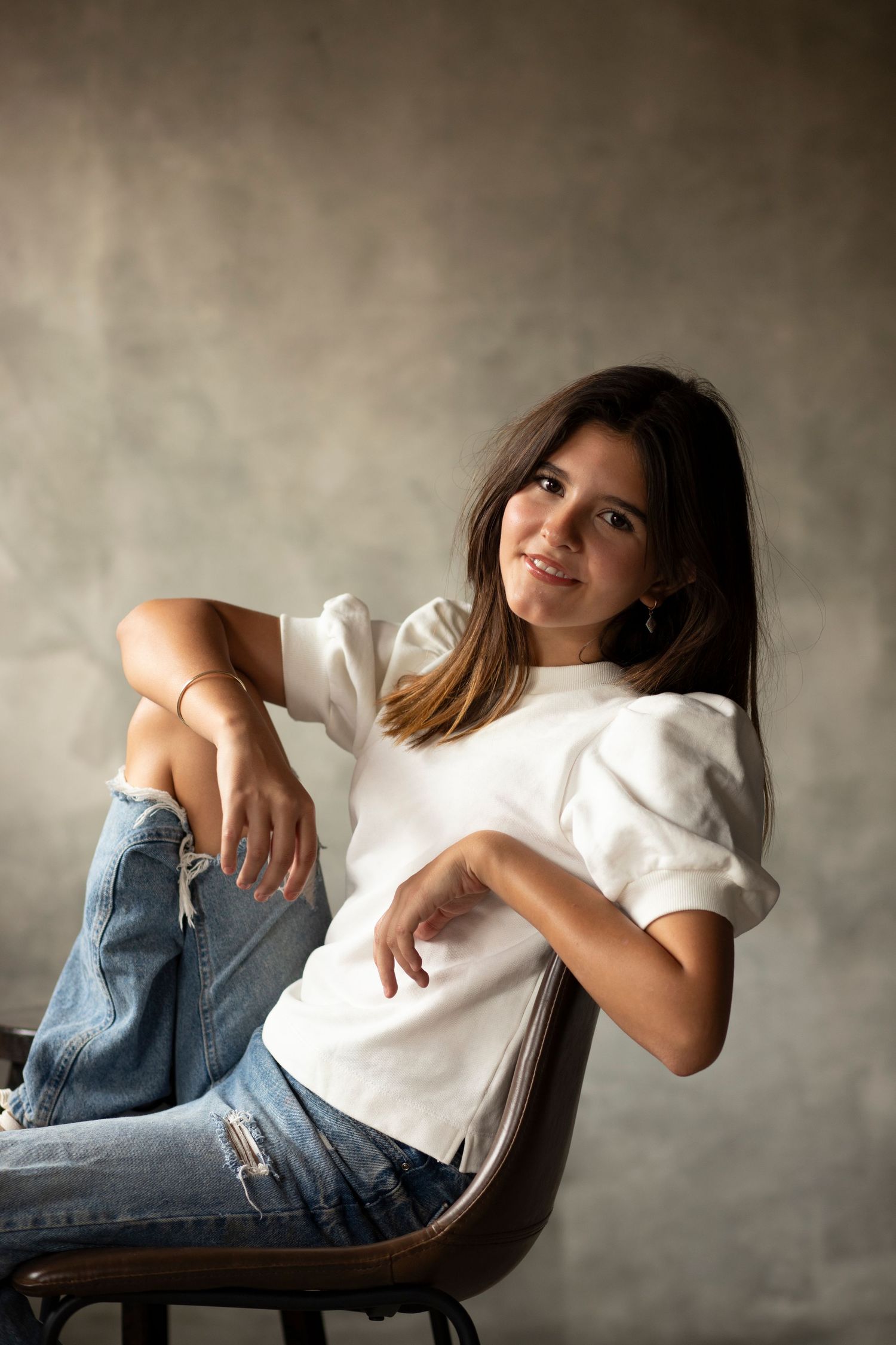 Casual studio portrait with relaxed pose on wooden chair against neutral backdrop.