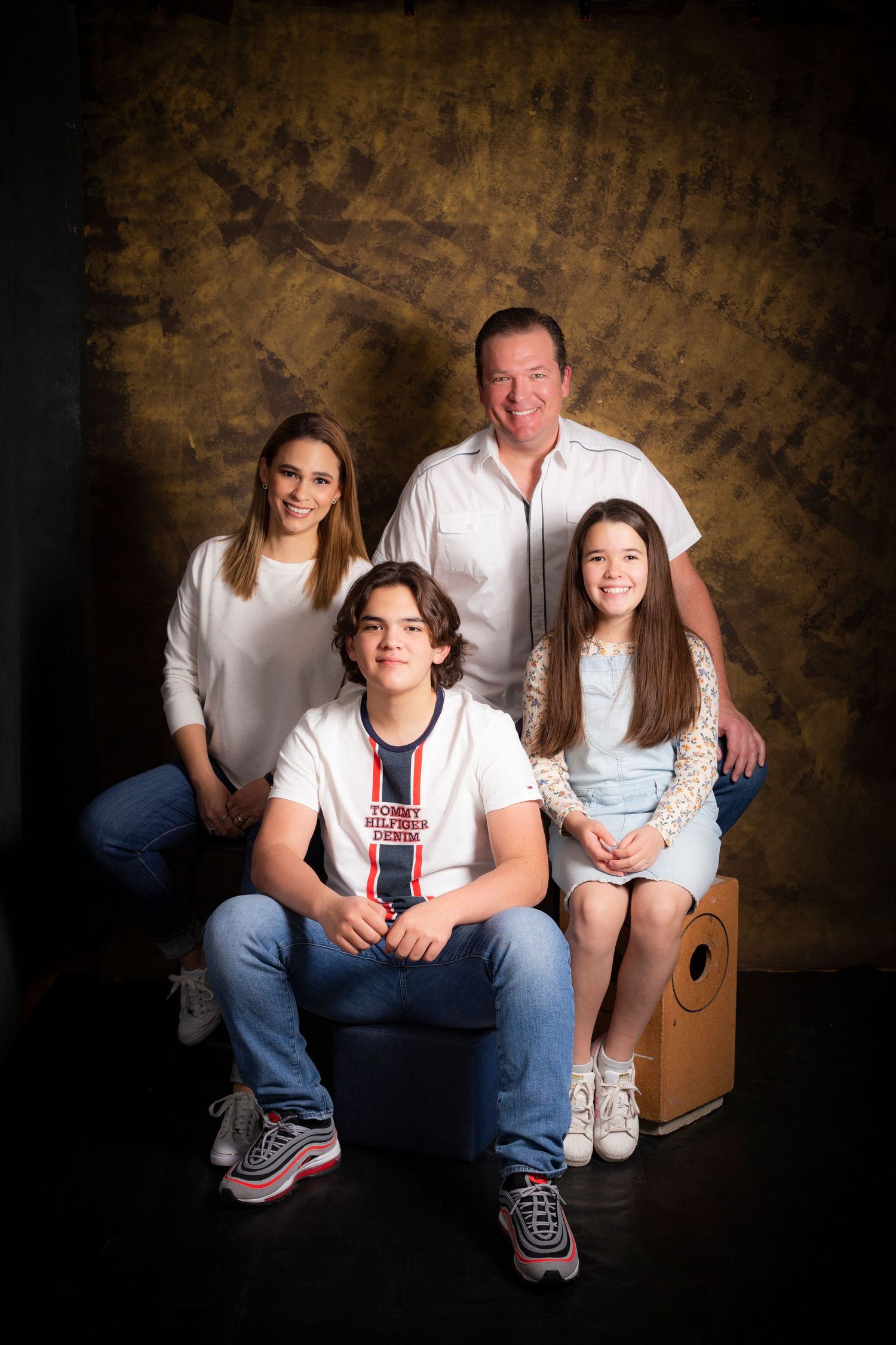 Studio portrait of a family of four in casual attire against a textured brown backdrop.