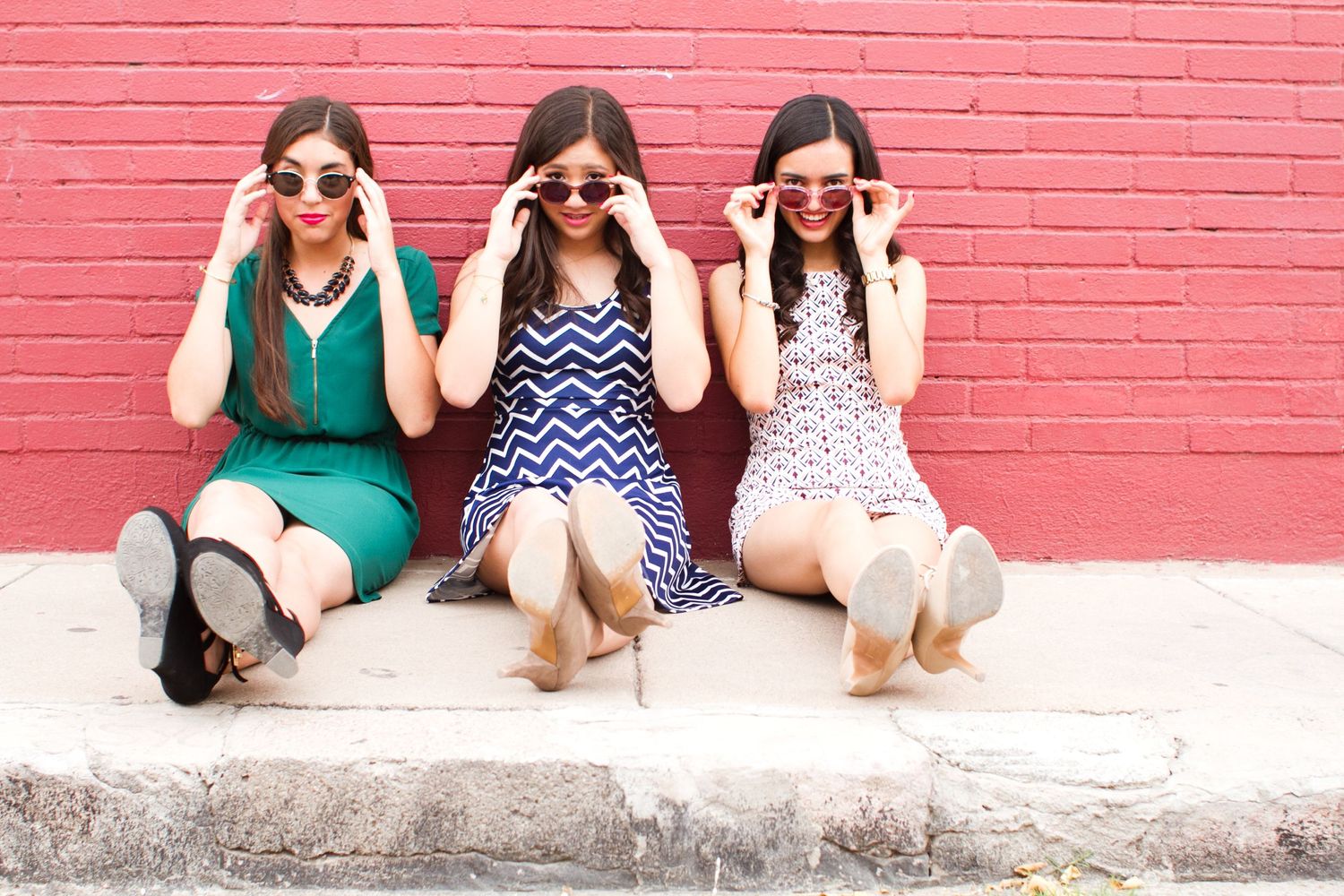 Three friends in summer dresses making silly faces while sitting against pink brick wall.