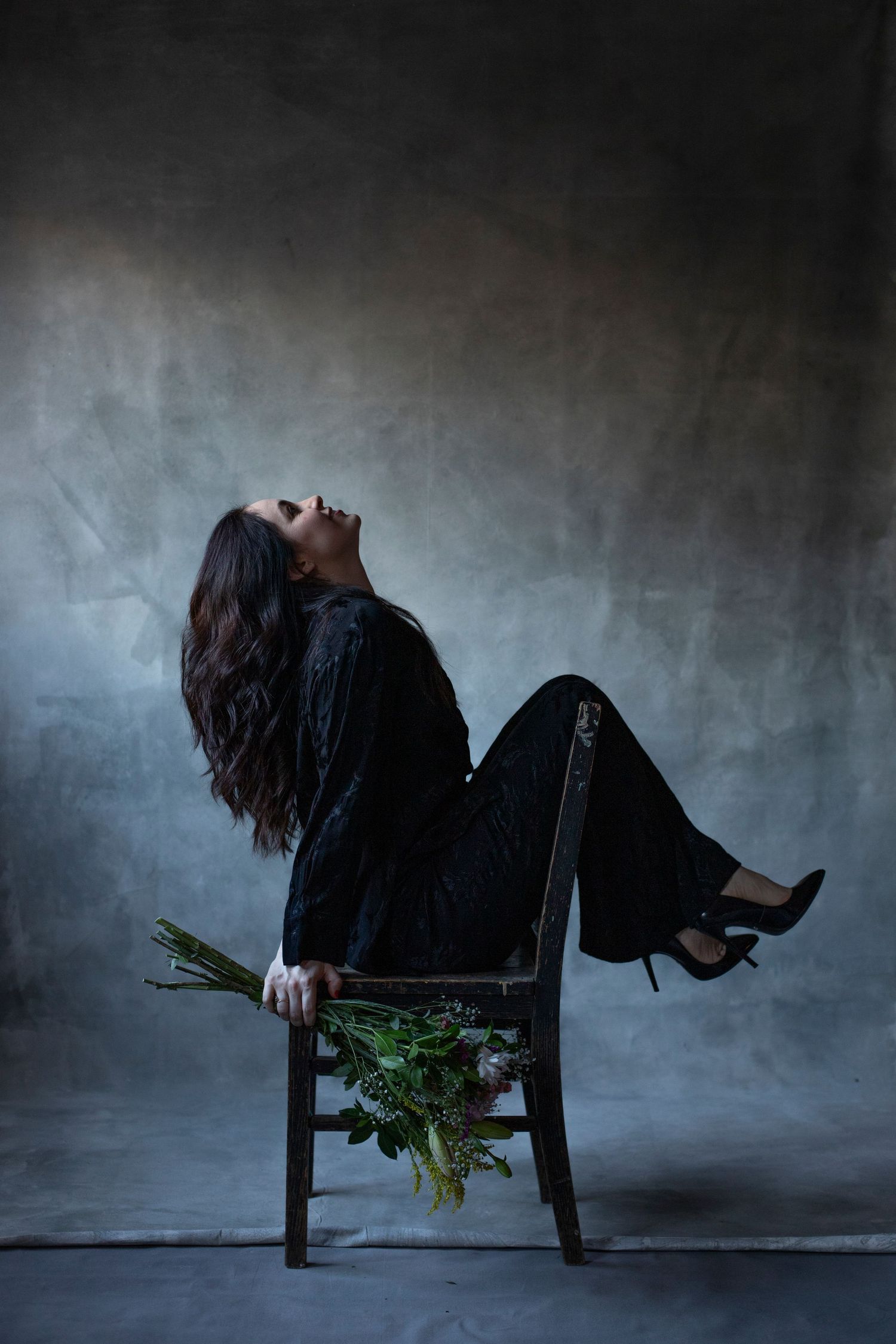 Dramatic portrait with figure reclining on chair holding flowers against dark background.