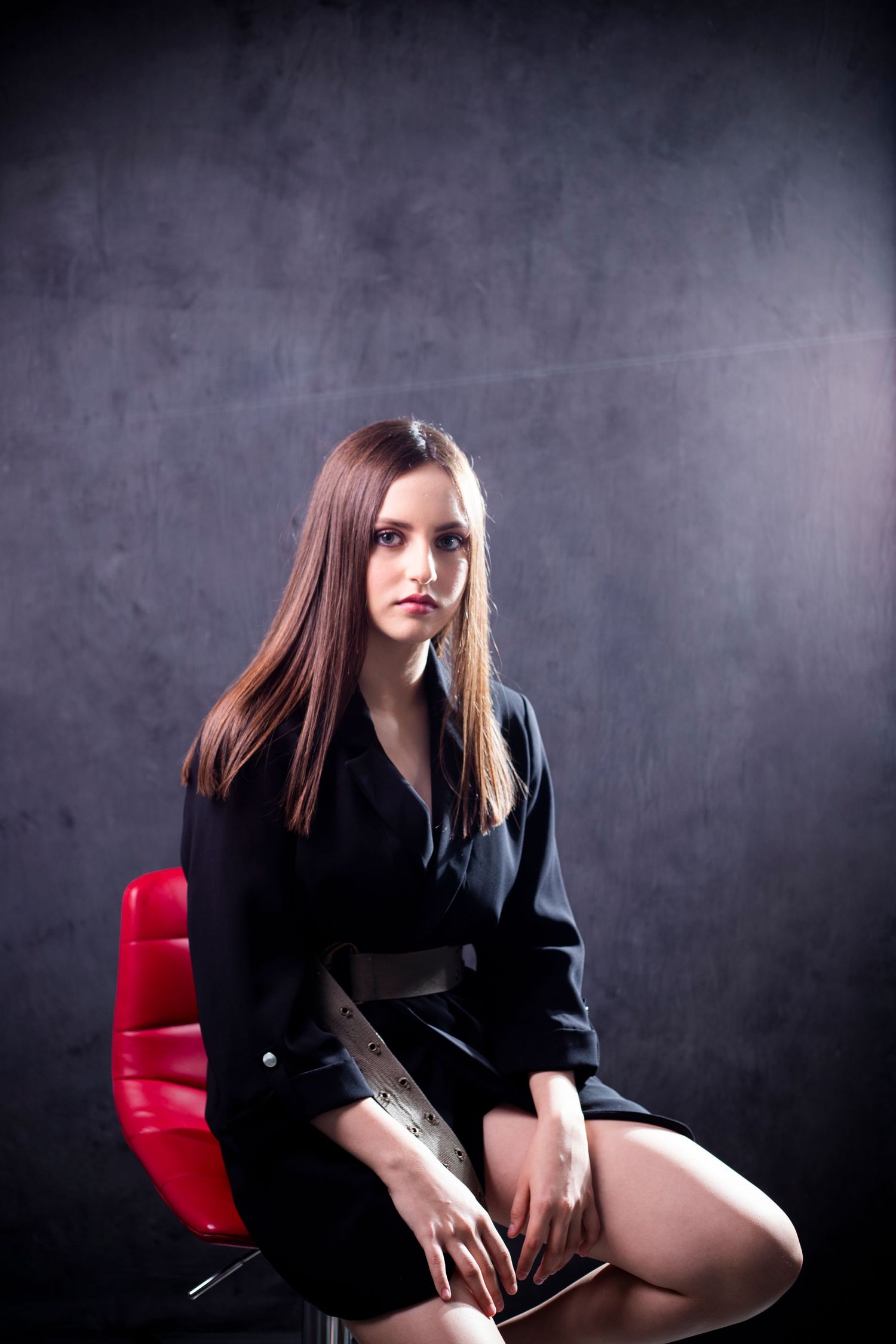 Studio portrait featuring a person in black attire seated on a red chair against a dark background.