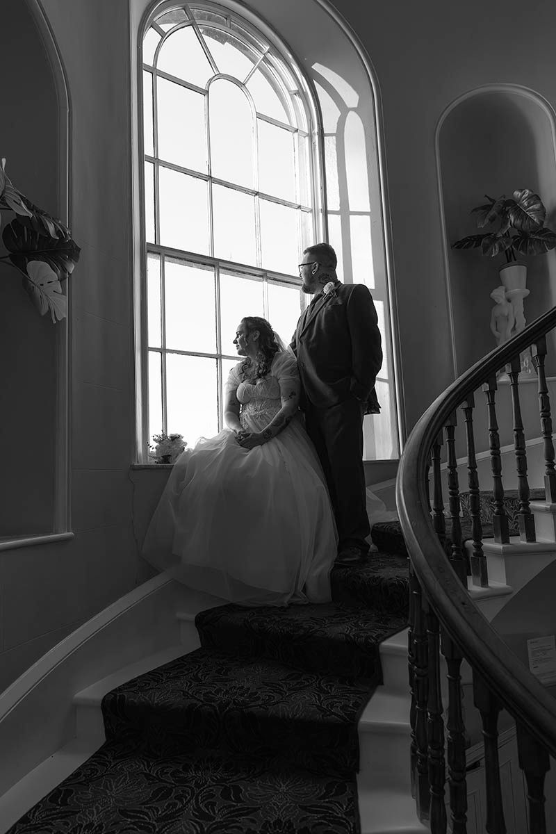 Black and white wedding photo of bride and groom standing on elegant staircase by arched window in grand venue.