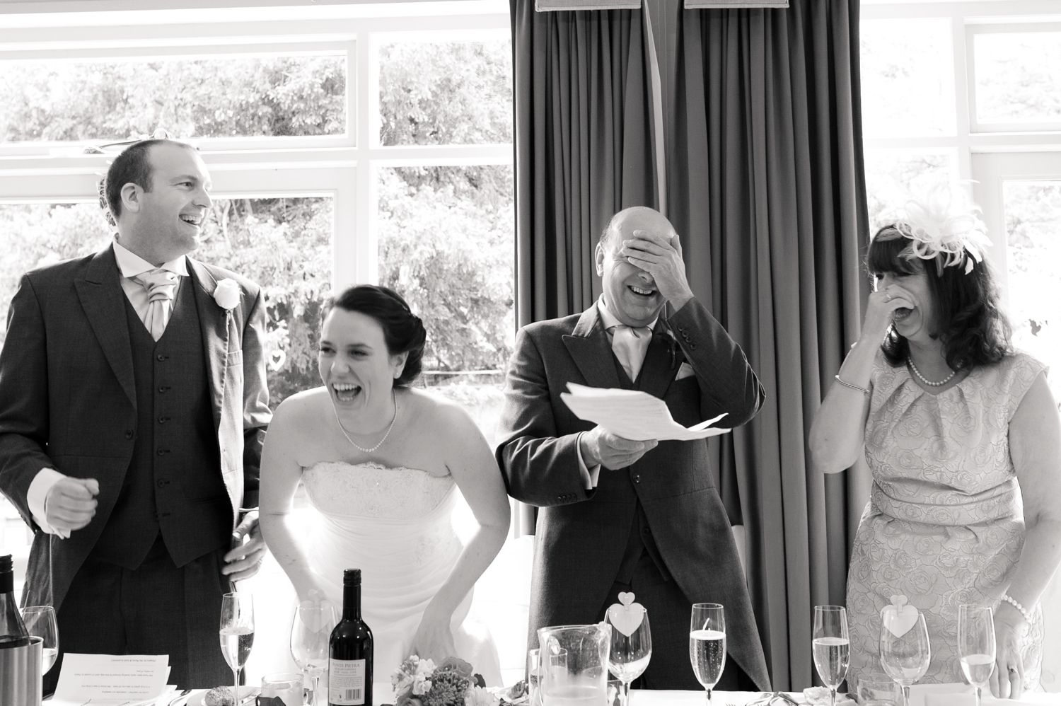 Black and white wedding photo of bride, groom, and guests laughing during a speech at a reception.